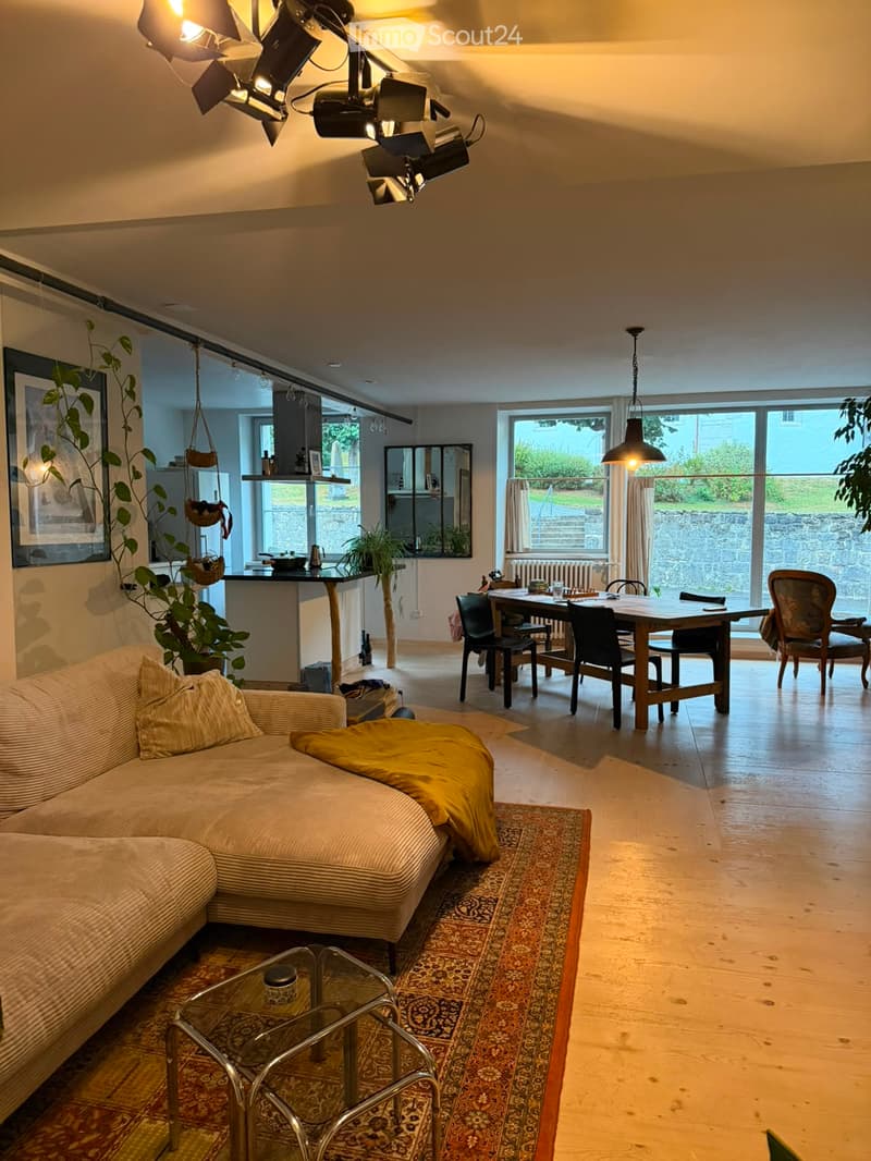 Open-plan living and dining area with a sofa, coffee table, dining table, chairs, pendant lights, and a large window overlooking a garden.