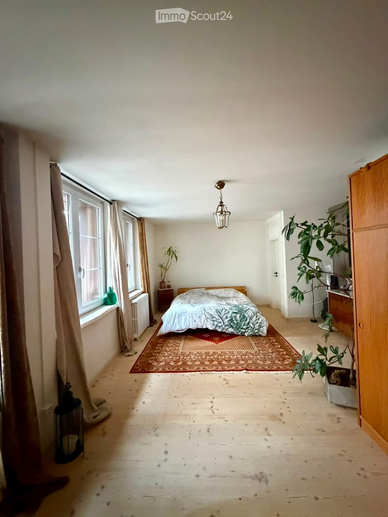 Cozy bedroom with wooden floor, large window with curtains, bed with patterned bedding, nightstand, and plants.