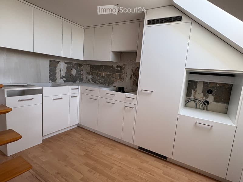 Modern kitchen with white cabinets, wooden floor, and exposed brick wall. Features a sink, stove, and a small wooden stool.