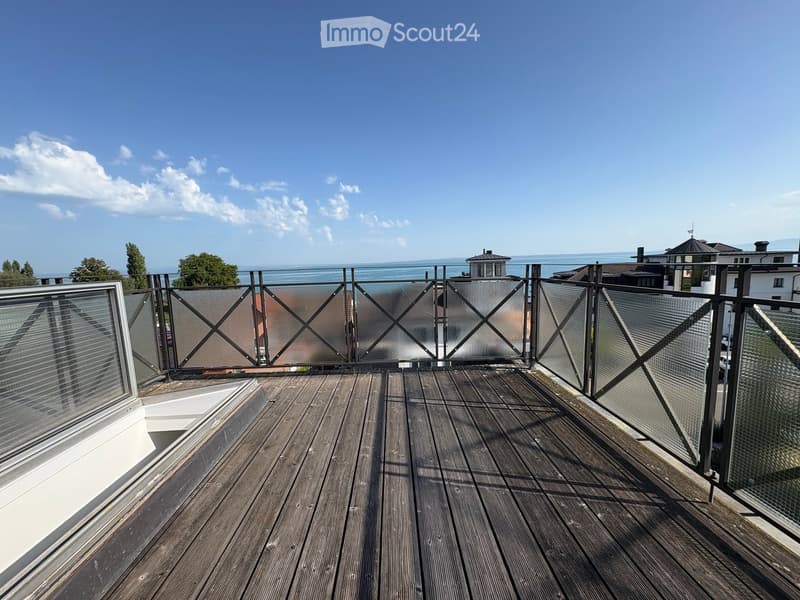 Wooden deck with modern glass railing, overlooking a body of water and distant buildings.