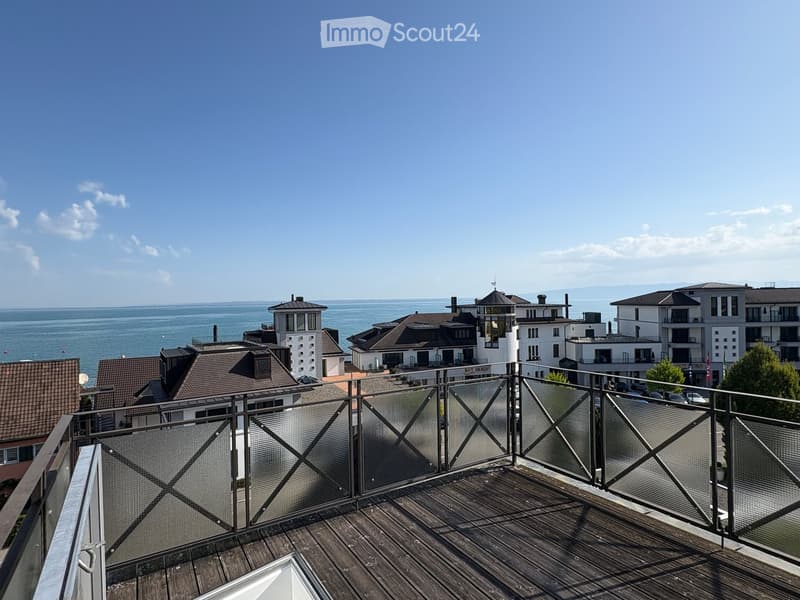 Wooden balcony with metal railing, overlooking a coastal town with buildings and the sea in the background.