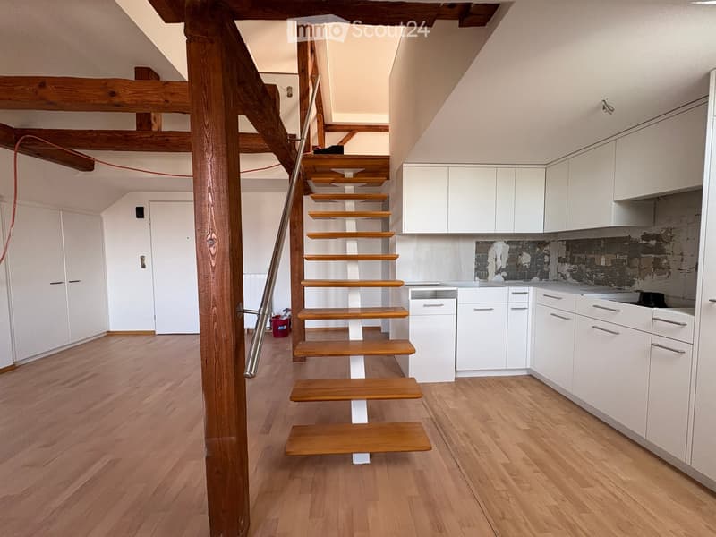 The image shows a modern, open-plan space with a wooden staircase leading to an upper level, white kitchen cabinets, and a kitchen counter area. The floor is made of light wooden planks, and the walls are painted white.
