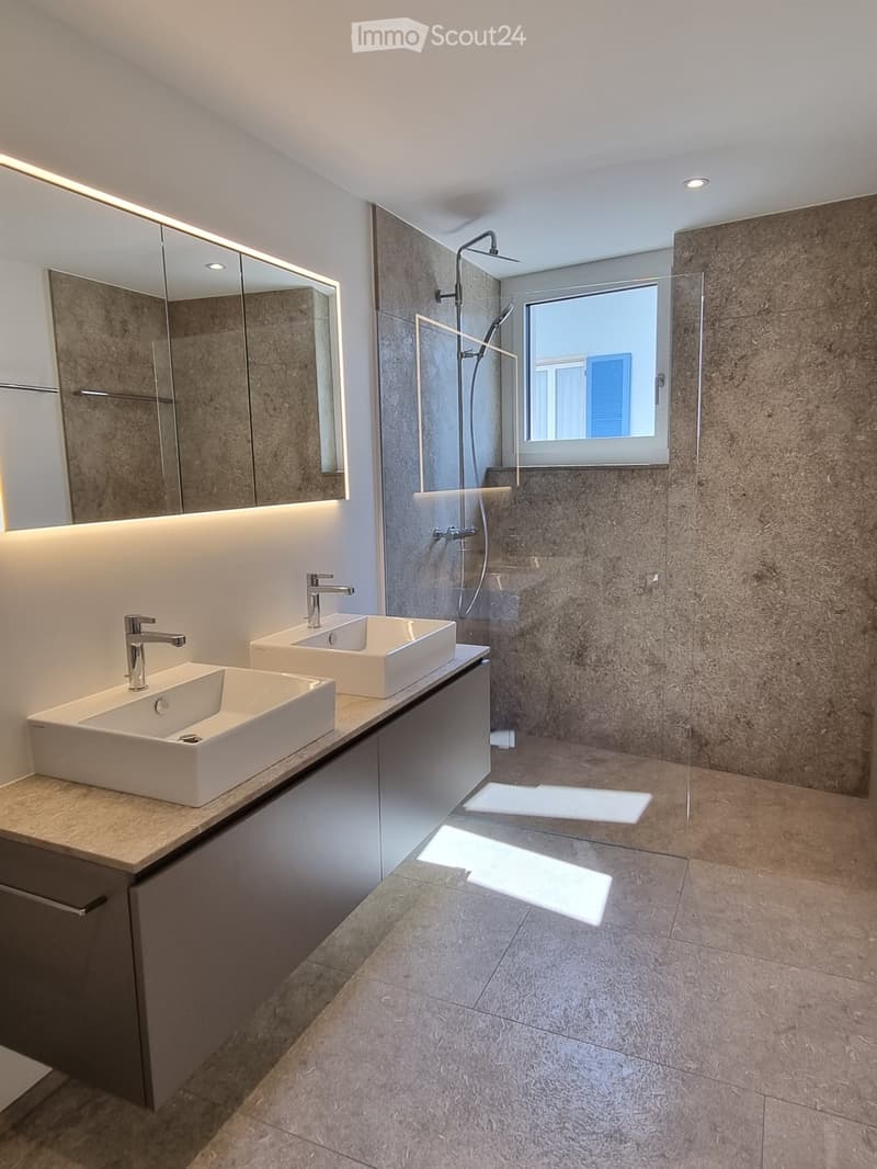 Modern bathroom with two white sinks, chrome faucets, large mirror with LED lighting, and a glass-enclosed shower area with a window.