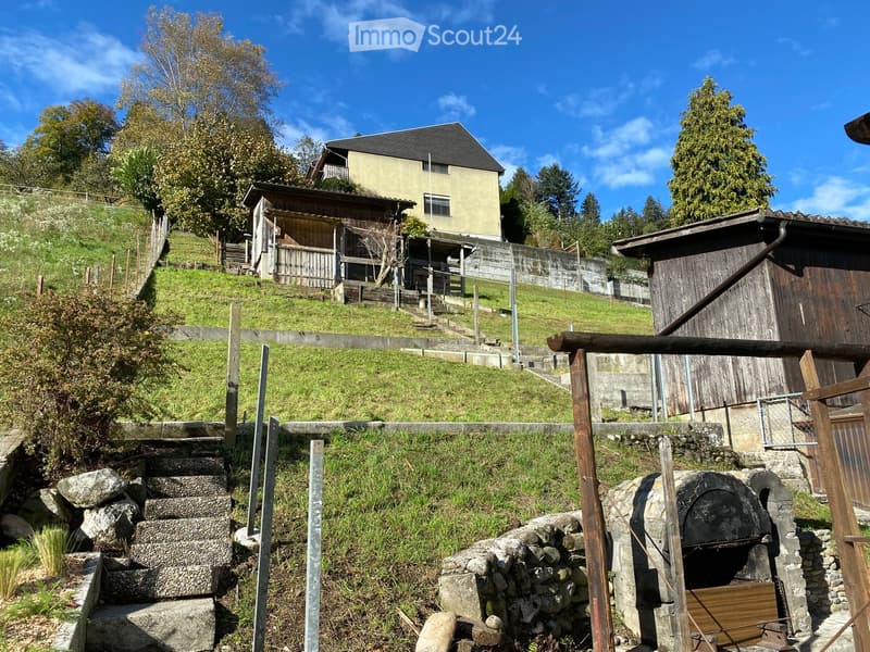 A rustic outdoor area with a stone staircase leading up to a wooden deck, surrounded by greenery and a stone oven structure. The area includes a raised wooden platform and a small wooden shed.