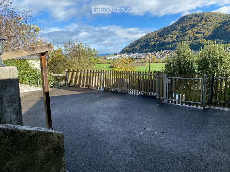 A paved outdoor area with a metal gate and wooden fence, surrounded by greenery and a scenic mountain view in the background.