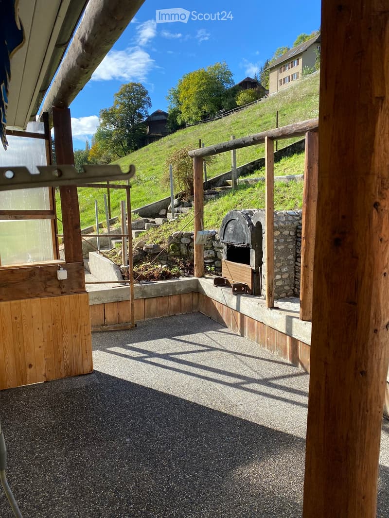 Outdoor patio area with gravel flooring, wooden railings, stone oven, and surrounding greenery.