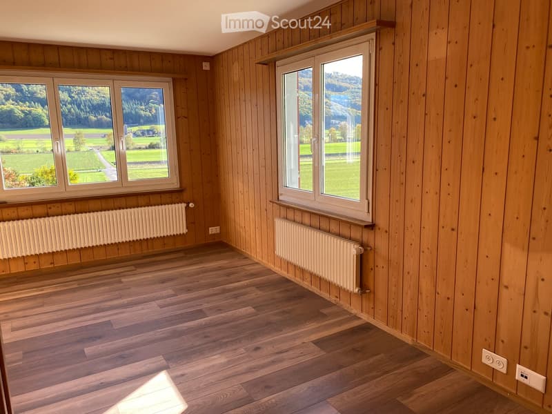 Empty room with wooden walls and flooring, large windows offering a scenic view, and radiators along the walls.
