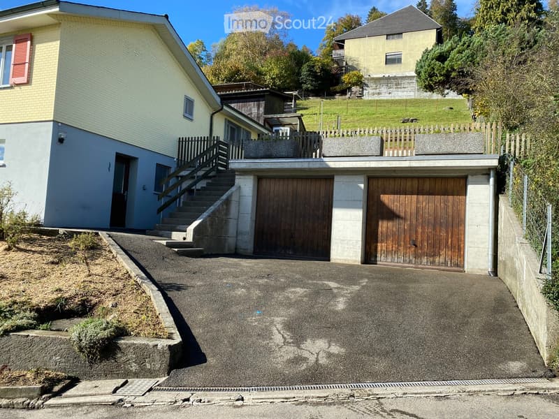 Two-story house with a garage, wooden garage doors, and a staircase leading to an upper level. The house has light yellow and white exterior walls, red shutters, and is situated on a sloped terrain with a concrete driveway.
