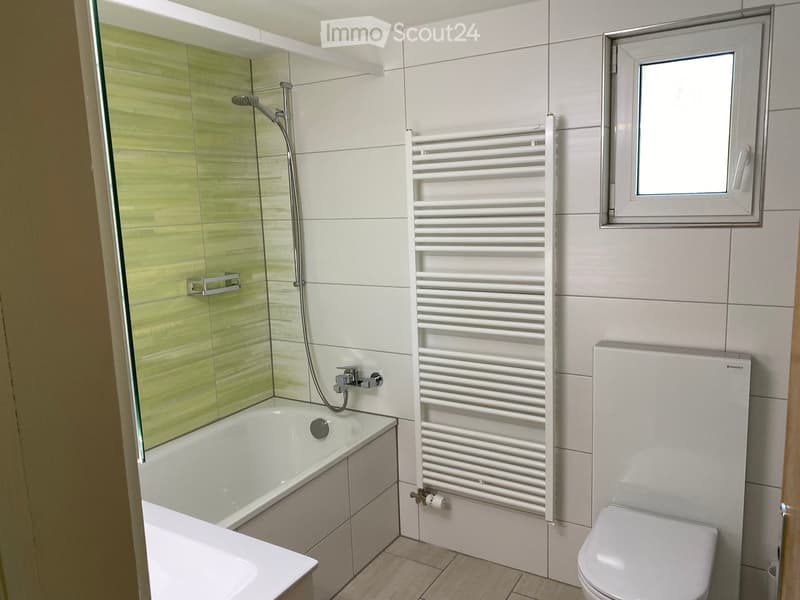 Modern bathroom with a bathtub, toilet, radiator, and a glass-enclosed shower area. The walls are tiled in white and green, and there is a window with a white frame.