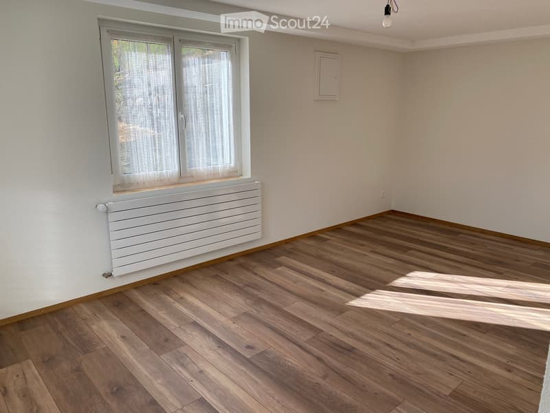 Empty room with wooden flooring, white walls, a window with white curtains, and a white radiator below the window.