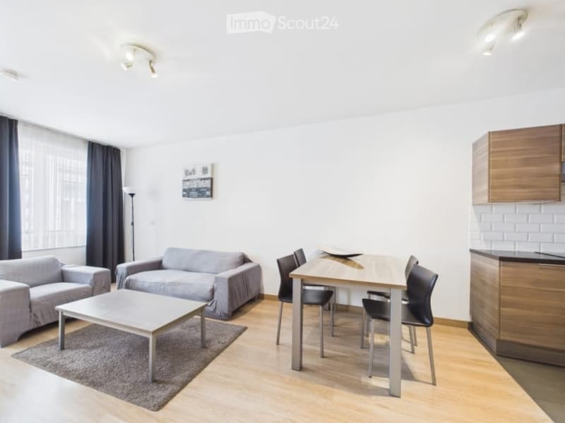 Modern, open-plan living and dining area with light wood flooring, white walls, grey sofa, coffee table, dining table with chairs, and kitchen cabinetry.