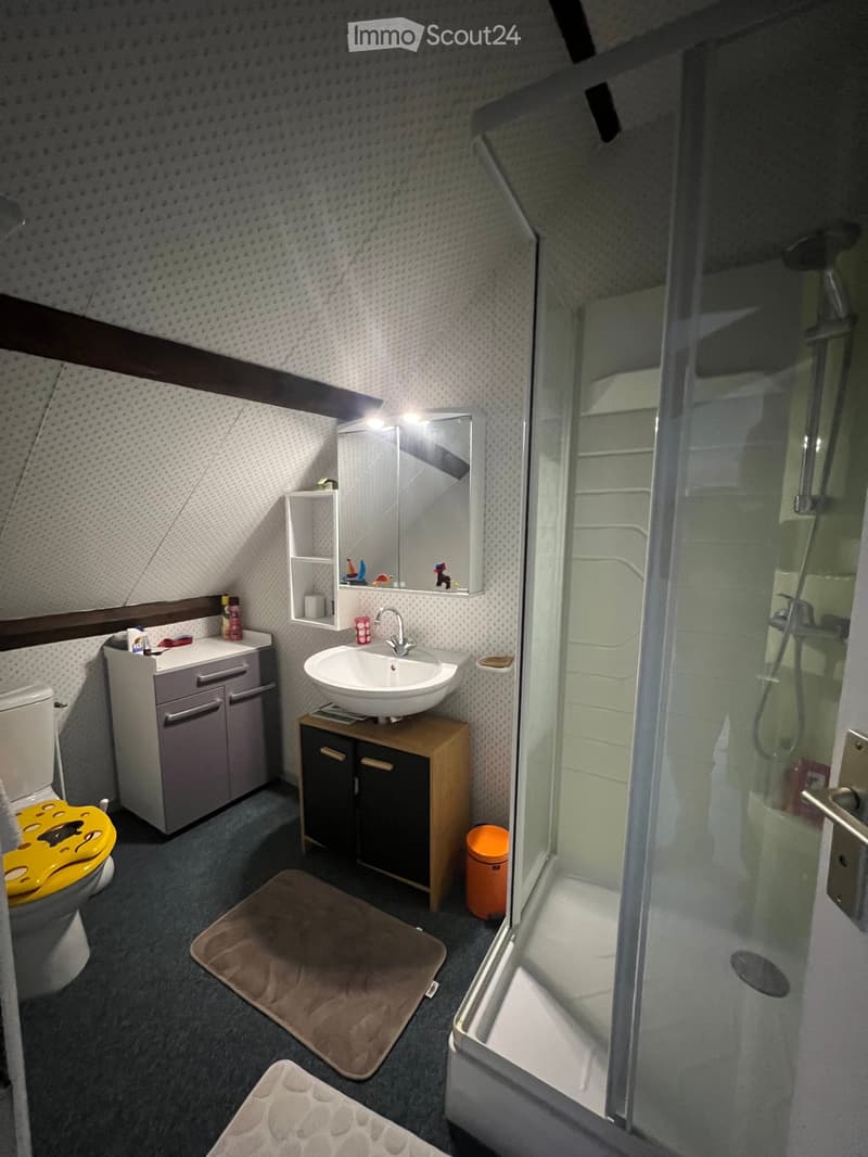 Cozy attic bathroom with a toilet, sink, mirror, shower, and small wooden cabinet. Features a slanted ceiling with exposed wooden beams and a small shelf above the sink.
