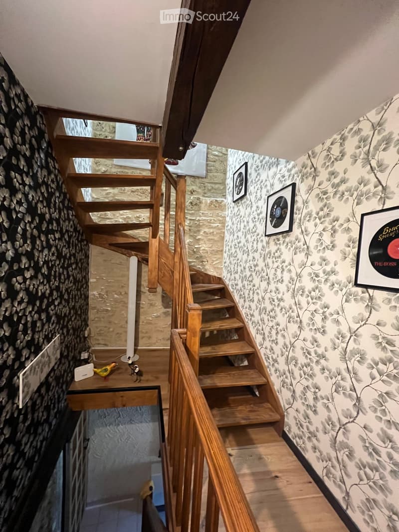 Wooden staircase with a wooden railing, patterned wallpaper, framed pictures, small decorative items on a shelf, and a small table with a bird figurine and a white device.