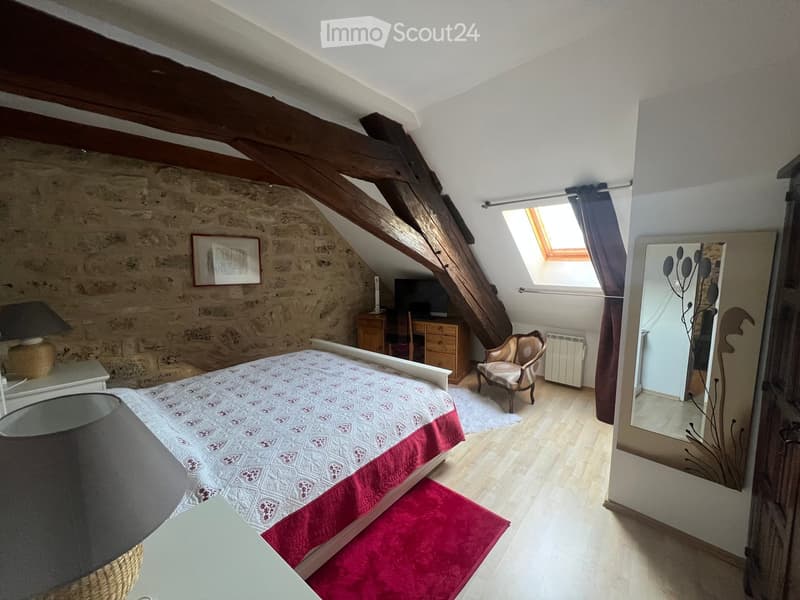 Cozy attic bedroom with exposed wooden beams, stone walls, a bed with patterned bedding, a wooden desk, a chair, a mirror, and a skylight window.