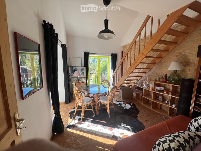 A cozy living room with a sofa, a glass dining table with chairs, a wooden staircase, a shelf with decorative items, and a large mirror on the wall.