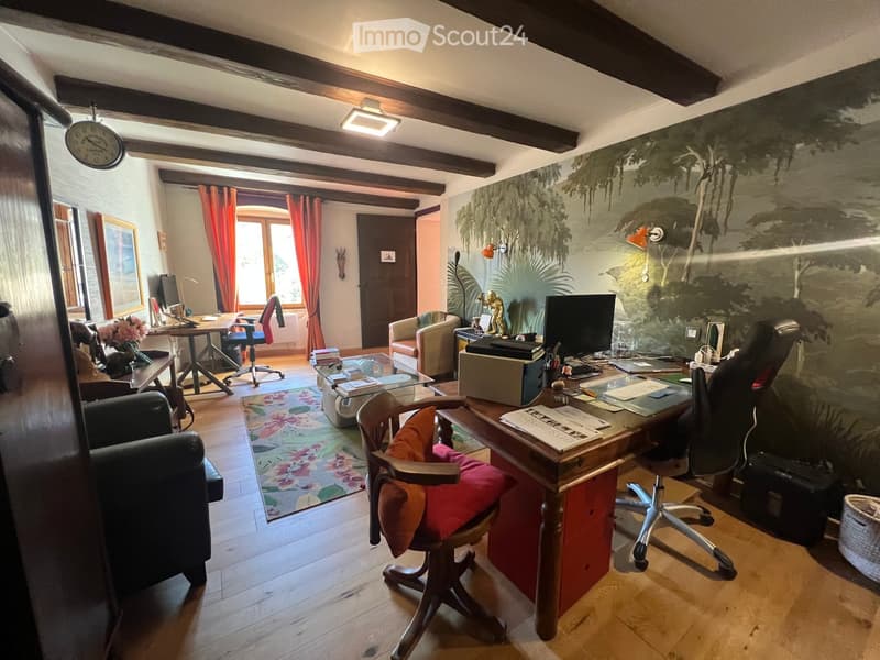 A well-furnished room with wooden floors, exposed wooden beams, a large desk with a computer, a comfortable sofa, a small table, and a colorful rug.