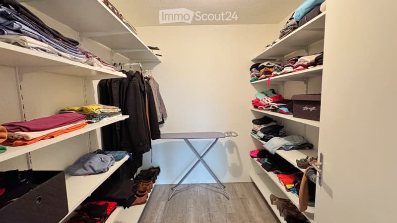 A well-organized closet with multiple shelves filled with folded clothes, hanging garments, and shoes. An ironing board is positioned in the center.