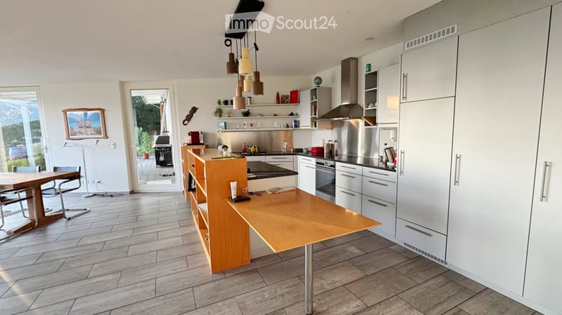 Modern kitchen with white cabinets, stainless steel appliances, a central island, and a dining area. Features pendant lights, a view through large windows, and a dining table with chairs.