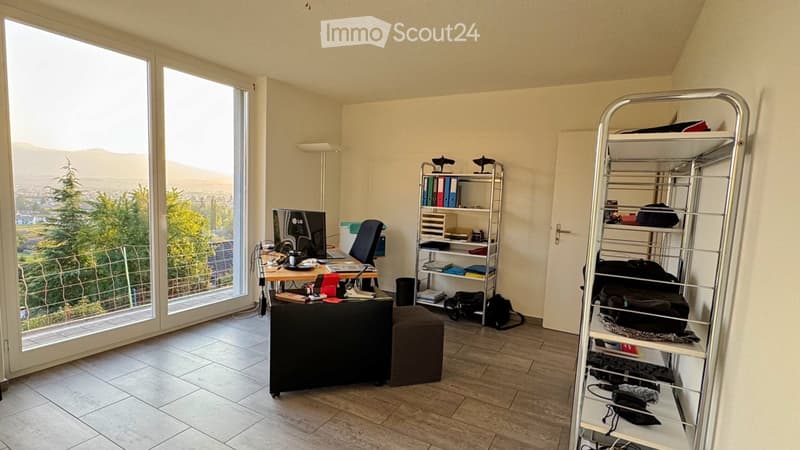 A well-lit room with large windows offering a scenic view, furnished with a desk, chair, computer monitor, stool, metal shelving unit, and various personal items.
