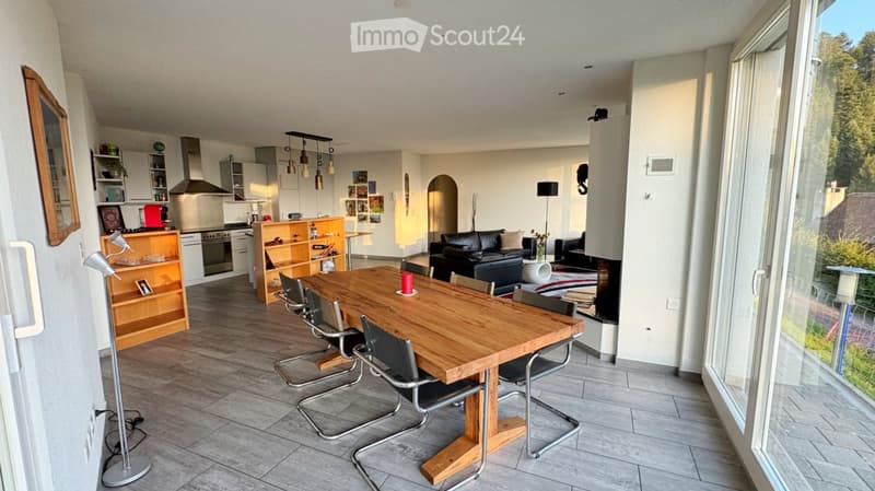 Open-plan living and dining area with a wooden dining table, chairs, a black leather sofa, a kitchen area with cabinets and appliances, and a floor lamp near the window.