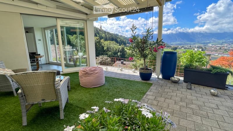 A modern balcony with a green artificial turf area, wicker chairs, a pink bean bag, a small table, and potted plants, offering a scenic mountain view.