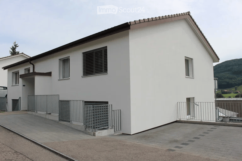 Single-story white house with a flat roof, multiple windows, and a small covered entrance area. The house is surrounded by a paved area with metal railings.