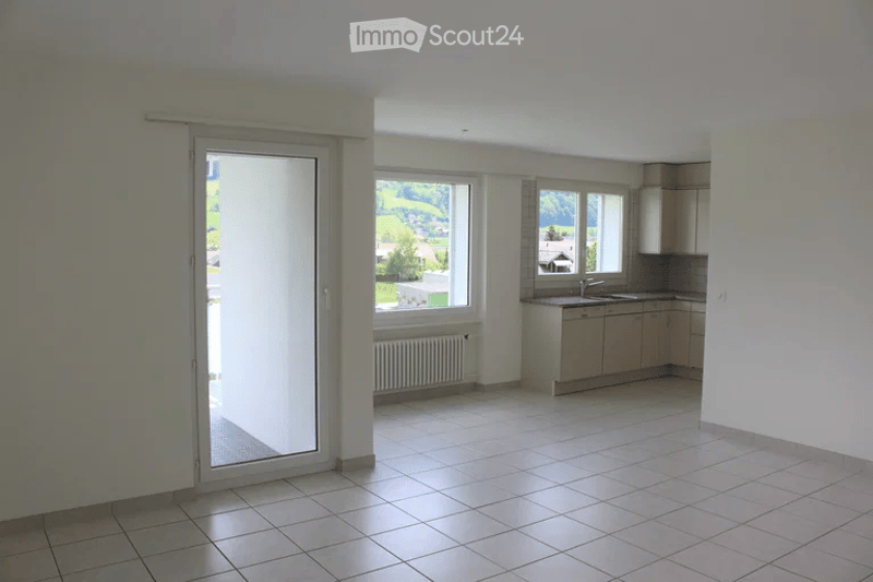 Bright, unfurnished room with large windows offering a scenic view, tiled floor, and a small kitchen area with cabinets and sink.
