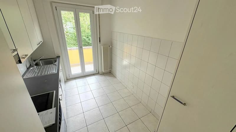 Modern kitchen with white tiled walls, white cabinets, a sink, and an oven. A sliding glass door leads to a balcony.