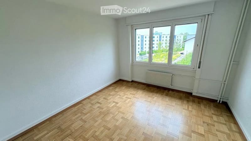 Empty room with wooden herringbone flooring, white walls, and large windows showing a view of buildings and greenery outside.