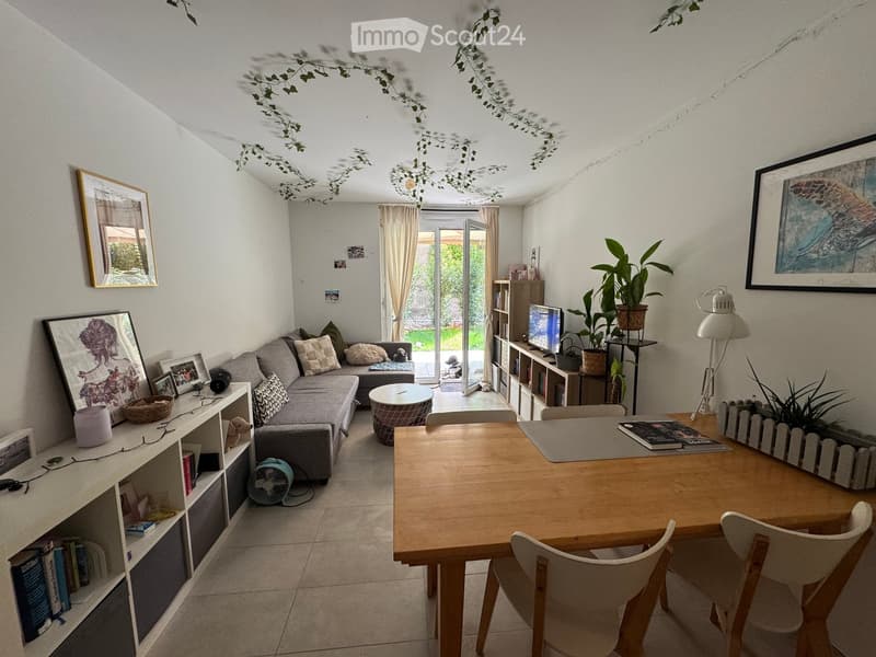 A modern living room with a grey sofa, coffee table, dining table with chairs, bookshelf, fan, lamp, picture frames, and plants.