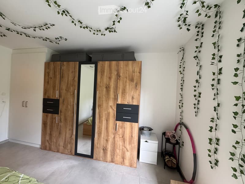 Bedroom with a large wooden wardrobe, mirror, small stool, fan, and shoes on a shelf. Green ivy garland decorates the ceiling and walls.