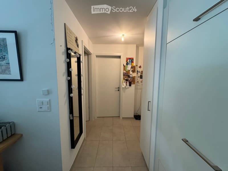 A narrow hallway with tiled flooring, white walls, and several doors leading to different rooms. A mirror with a string of lights is mounted on one wall, and a small shelf is visible on the left side.