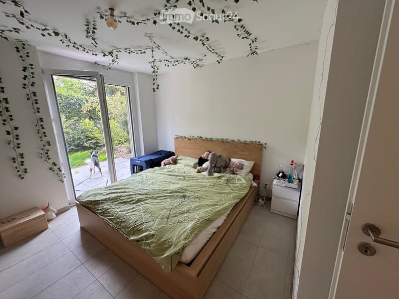Cozy bedroom with a wooden bed, green duvet, nightstand, and decorative items. A large window with a view of a garden and a dog is visible outside.