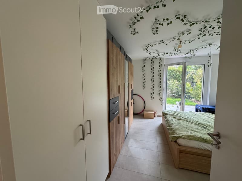 Bright bedroom with a wooden bed, wardrobe, and a large mirror. The room features a slanted ceiling with decorative ivy garlands and a sliding glass door leading to a garden.
