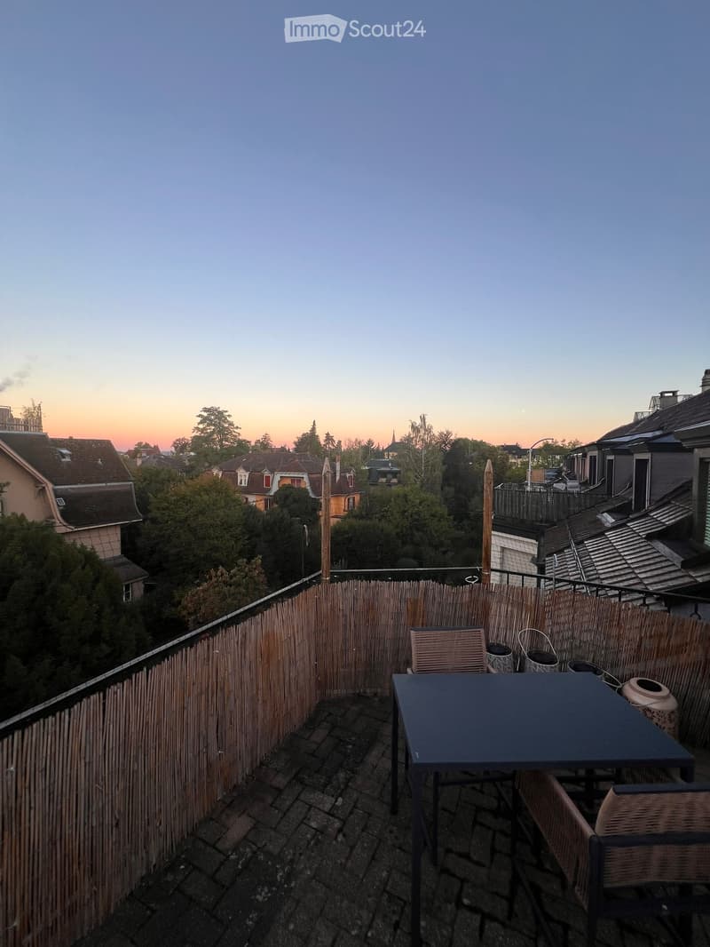 Balcony with a wooden fence, a table, and chairs, overlooking a cityscape at sunset.