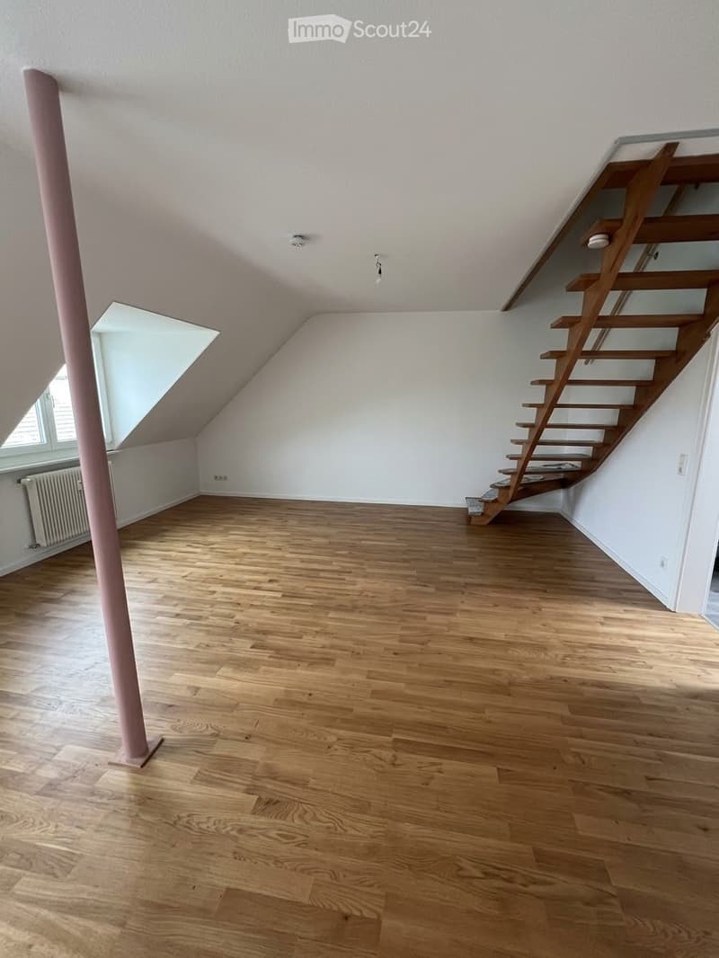 Empty attic room with wooden flooring, white walls, and a staircase leading upwards. Features a skylight window and a pink support column.