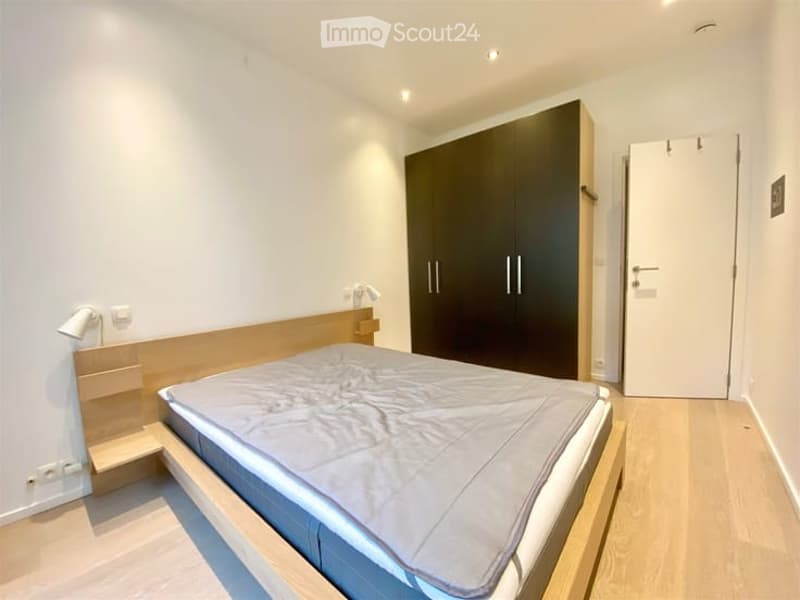 Modern bedroom with a wooden bed frame, grey bedding, large dark wardrobe, and a wall-mounted lamp. Light wooden flooring and white walls.