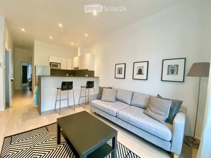 Modern, open-concept living and dining area with a grey sofa, coffee table, and two stool chairs at a kitchen island. The kitchen features white cabinets and stainless steel appliances, including a microwave and refrigerator. A floor lamp and framed pictu
