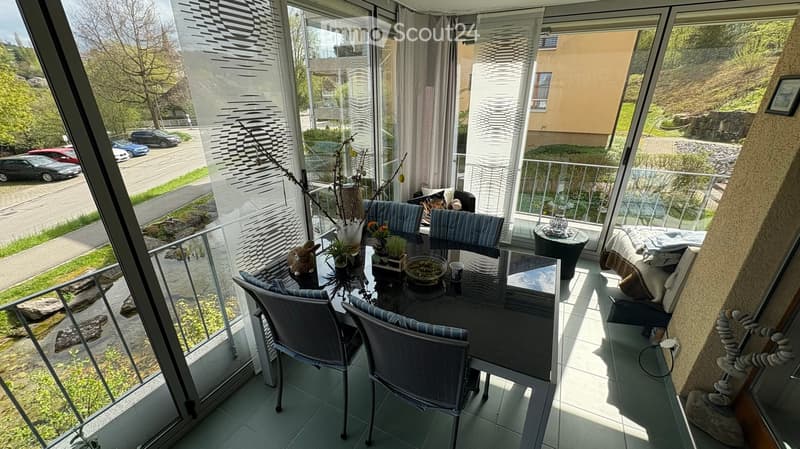 Balcony with a dining table, chairs, and decorative items, overlooking a street with parked cars and greenery.