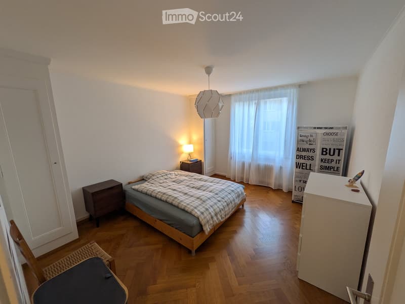 Cozy bedroom with wooden flooring, a single bed with plaid bedding, a nightstand with a lamp, a chair, and a mirror. The room features a large window with white curtains and decorative wall art.