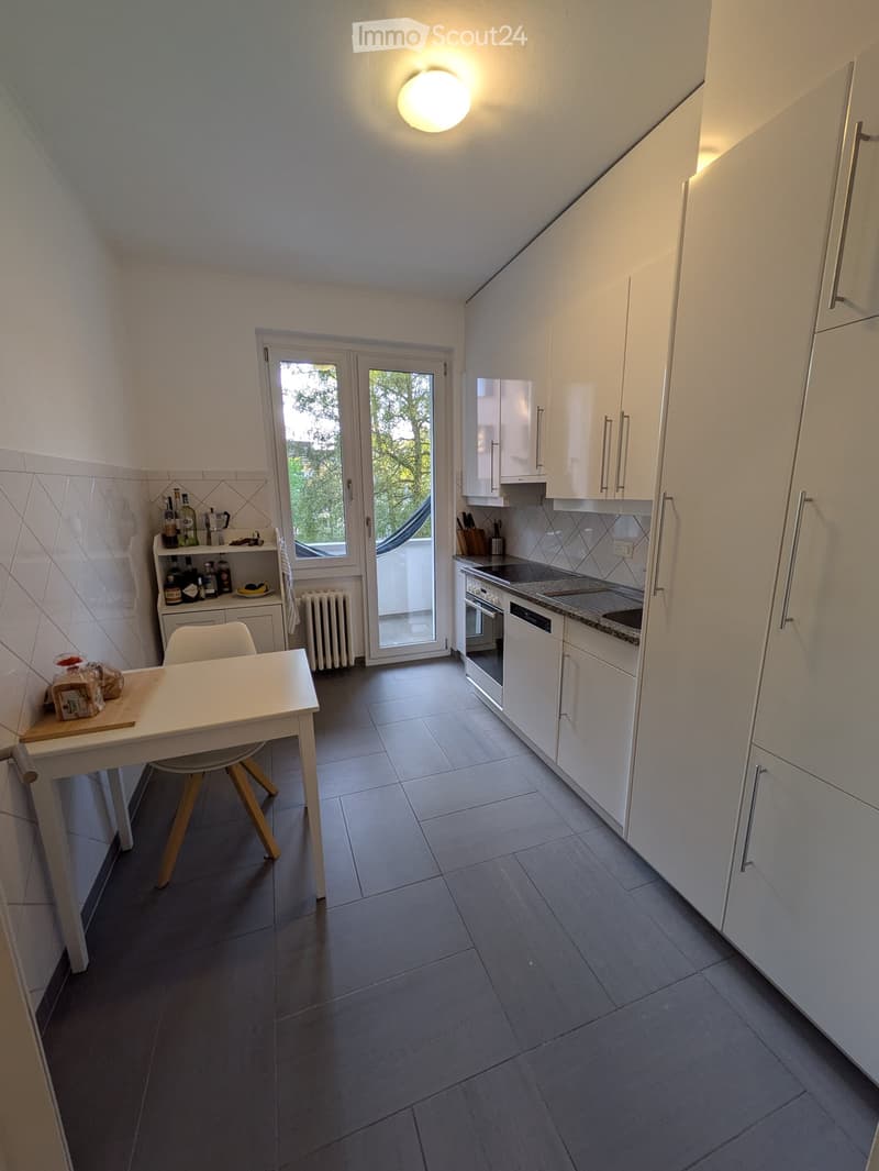 Modern kitchen with white cabinets, dark tiled floor, and a small dining area. Features include an oven, dishwasher, refrigerator, and a small table with a chair.