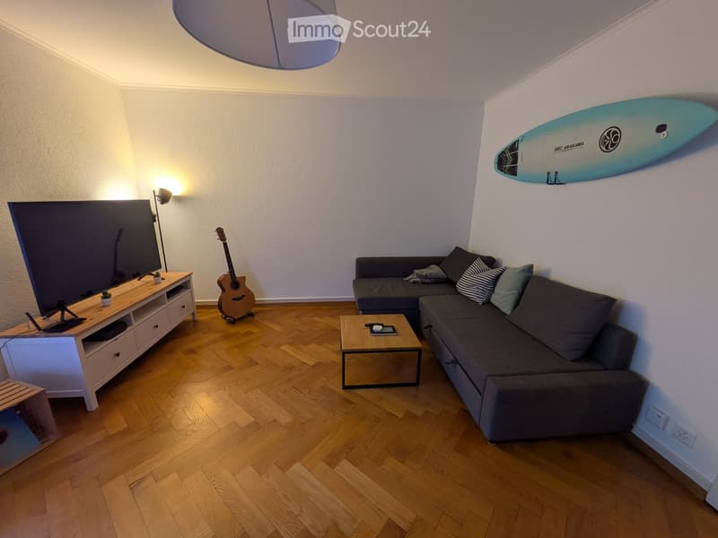 Modern living room with wooden flooring, grey sectional sofa, coffee table, TV stand, guitar, and a surfboard on the wall.