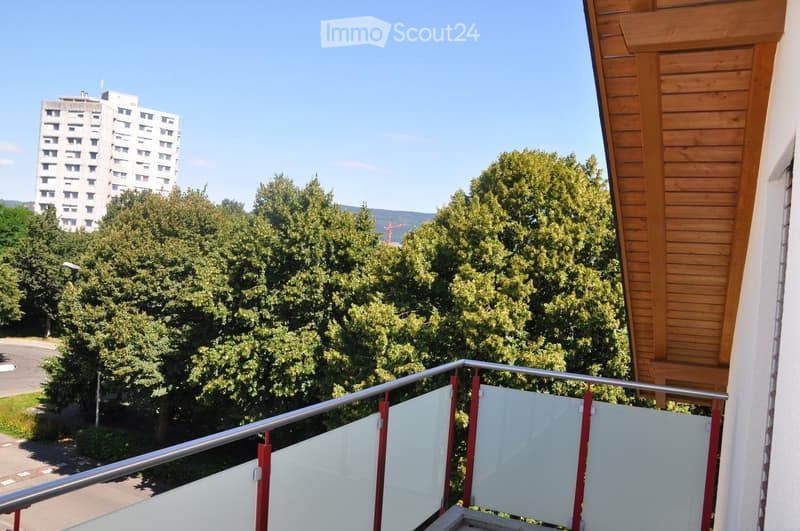 Balcony with a metal railing and glass panels, overlooking a green area with trees and a distant building.