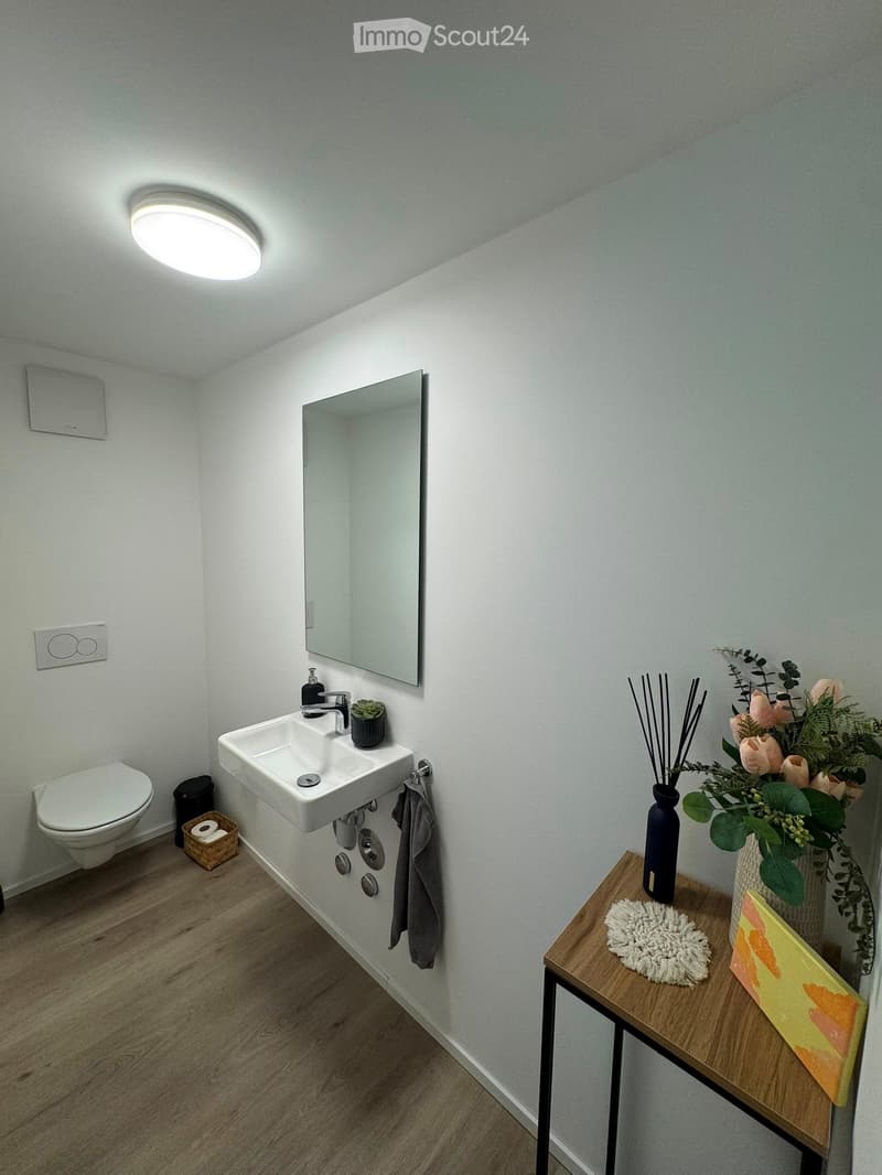 Modern bathroom with a white sink, chrome faucet, large mirror, toilet, and a small wooden table holding a vase with flowers, incense sticks, and a decorative pillow.