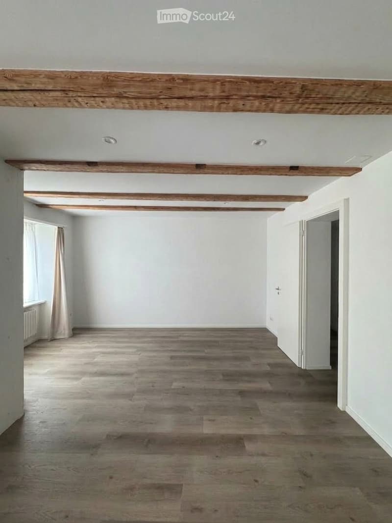 Empty room with wooden flooring, white walls, exposed wooden beams on the ceiling, and a window with a curtain on the left side.
