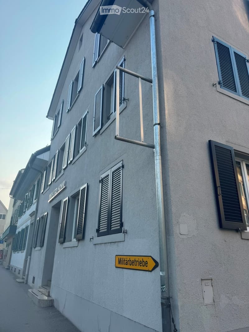 A multi-story building with grey walls, multiple windows with shutters, and a yellow sign reading 'Militärbetriebe'.