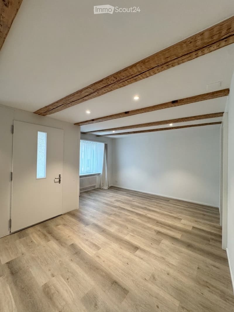 Empty room with light wooden flooring, white walls, exposed wooden beams on the ceiling, and a door leading to another room with a window and radiator.