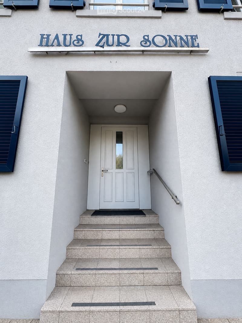 Entrance to a building named "HAUS ZUR SONNE" with a raised ground floor, featuring a staircase leading to a white door with a glass panel, flanked by dark blue shutters.