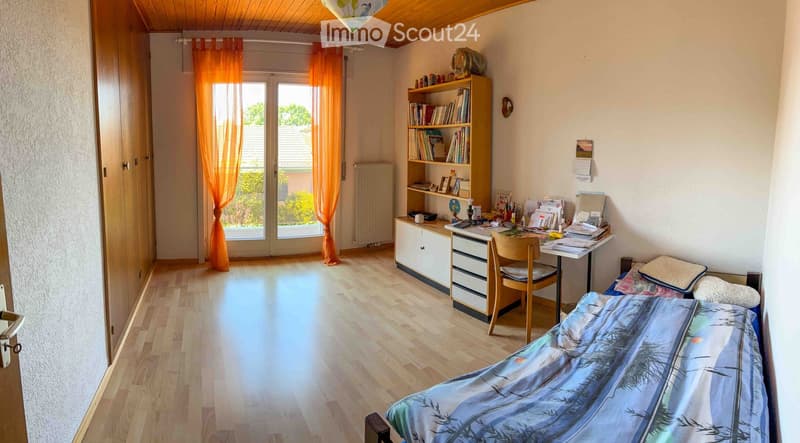 Cozy bedroom with wooden flooring, a bed with a blue and white duvet, a desk with a chair, a bookshelf, and a large window with orange curtains leading to a balcony.