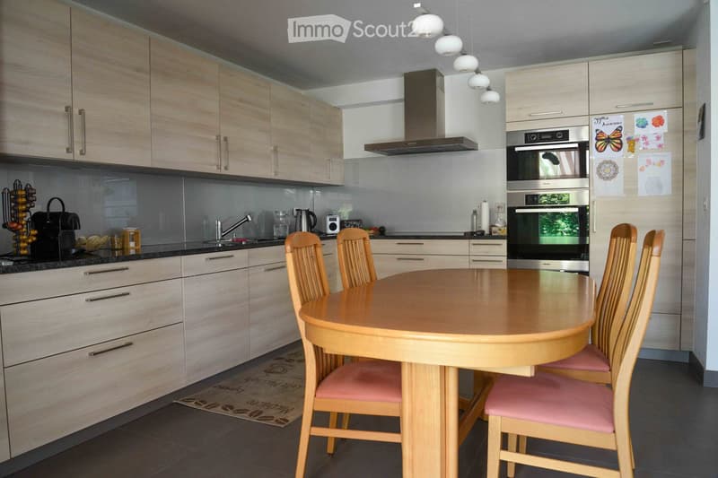 Modern kitchen with light wood cabinets, stainless steel appliances, a round wooden dining table with four chairs, and a hanging light fixture.